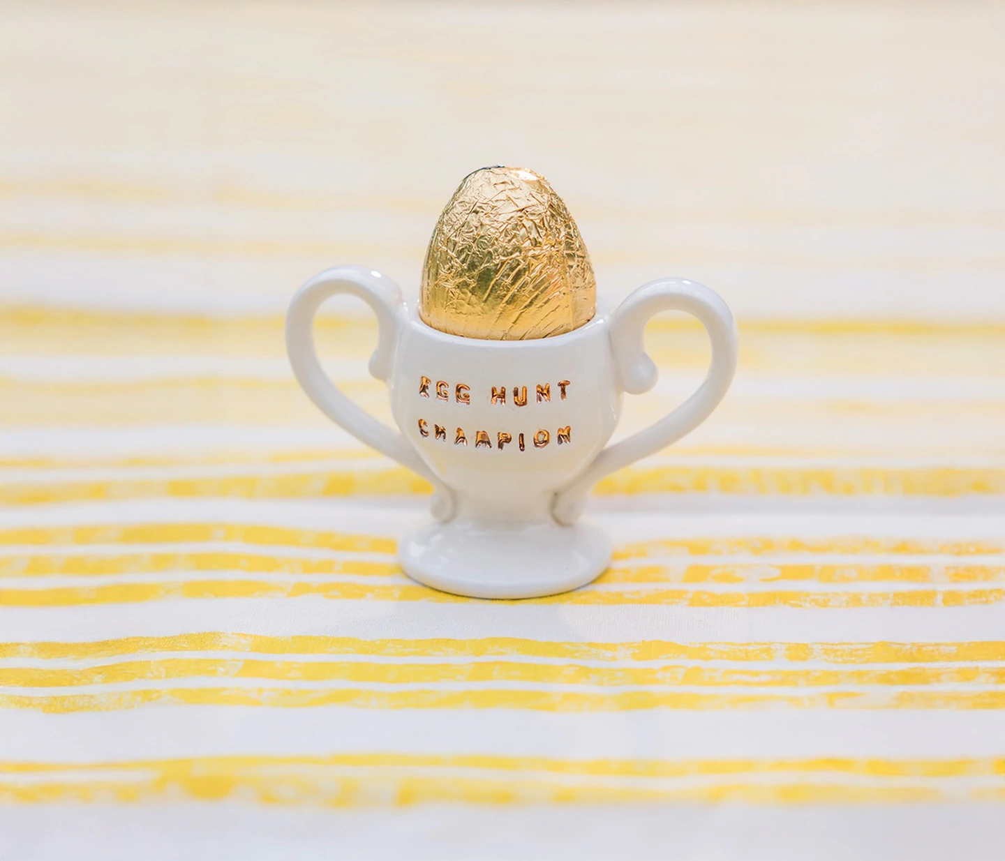 Egg hunt champion egg cup. A ceramic egg cup with the words 'Egg hunt champion' stamped in gold on a yellow tablecloth