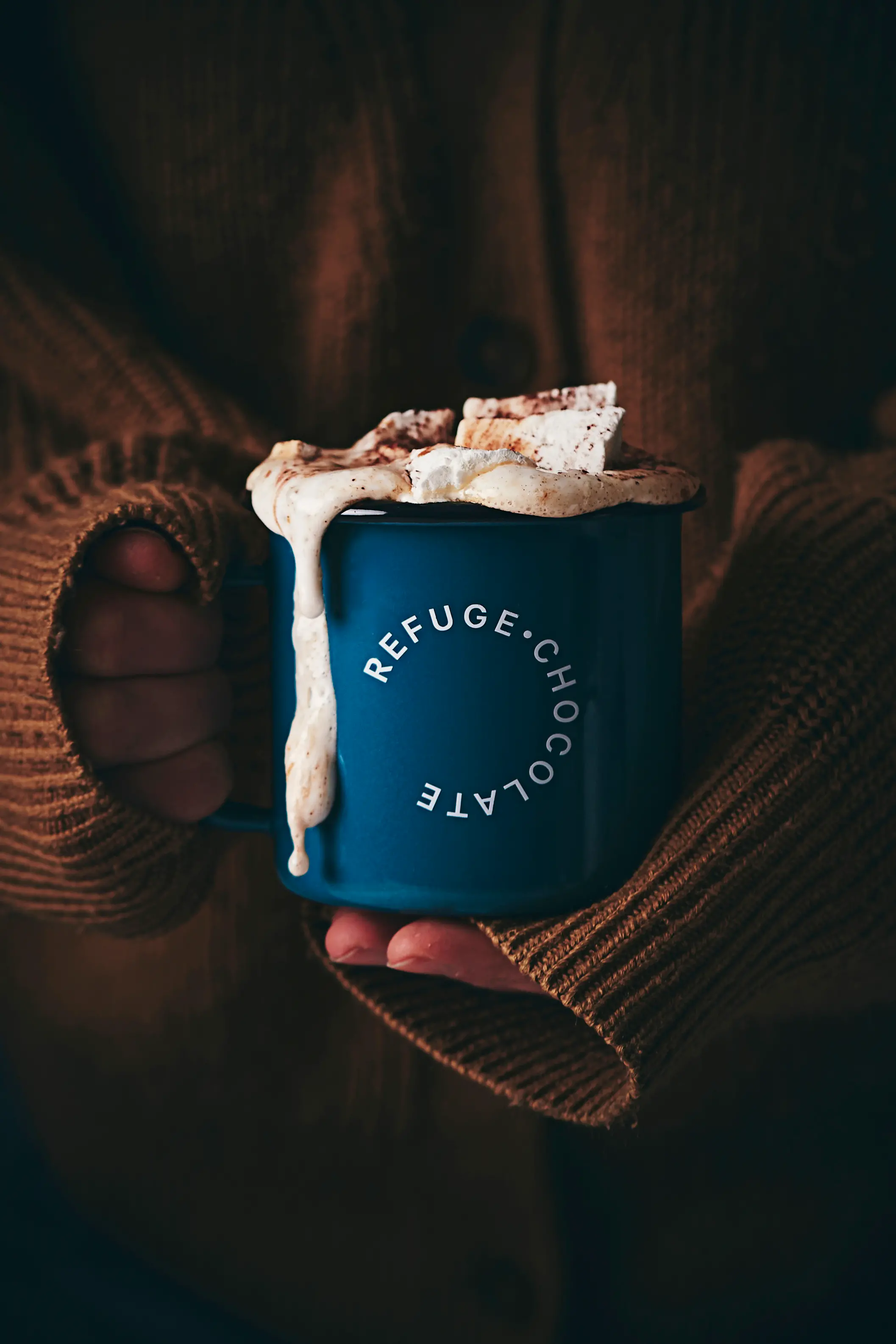 Branded Refuge Chocolate tin mug held in hands of cosy jumper. Melted marshmallows on top of the hot chcoolate.