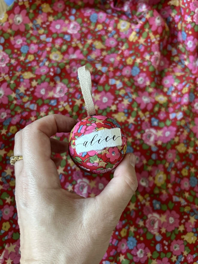 Mini Liberty fabric Christmas bauble with personalised Alice label held in front of a Liberty fabric backdrop 