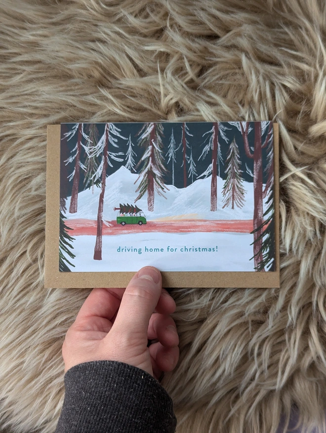 Driving home for Christmas card, shows a camper van carrying a Christmas tree through a winter forest.