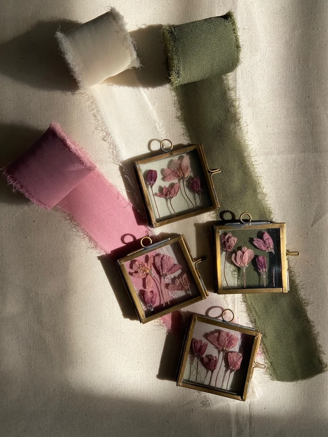Four square brass frames with small pink pressed flowers laid over pink, ivory and green chiffon ribbon