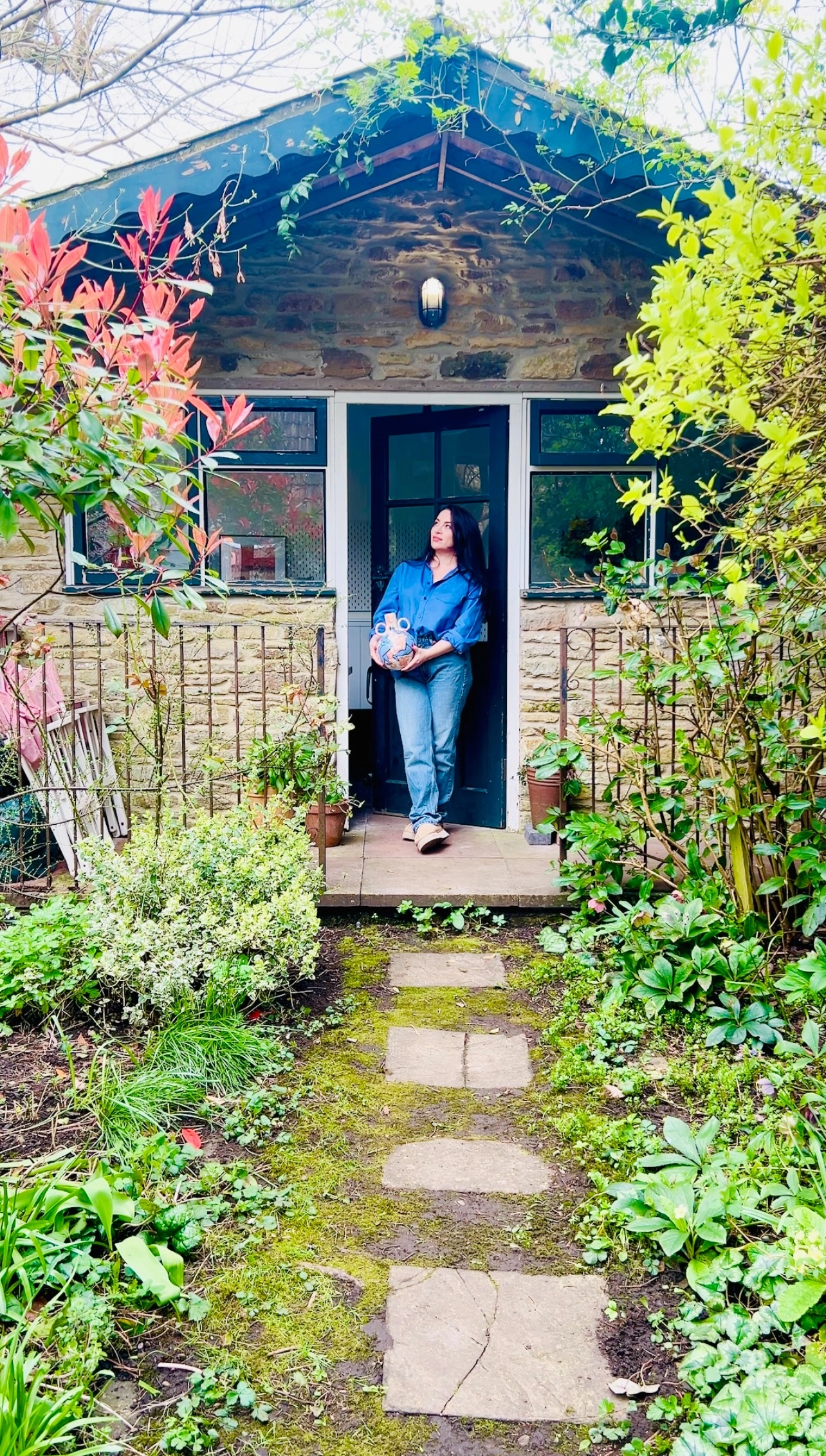 Woman holding a vase stands in the doorway of a stone garden studio, surrounded by lush green plants and a garden path.