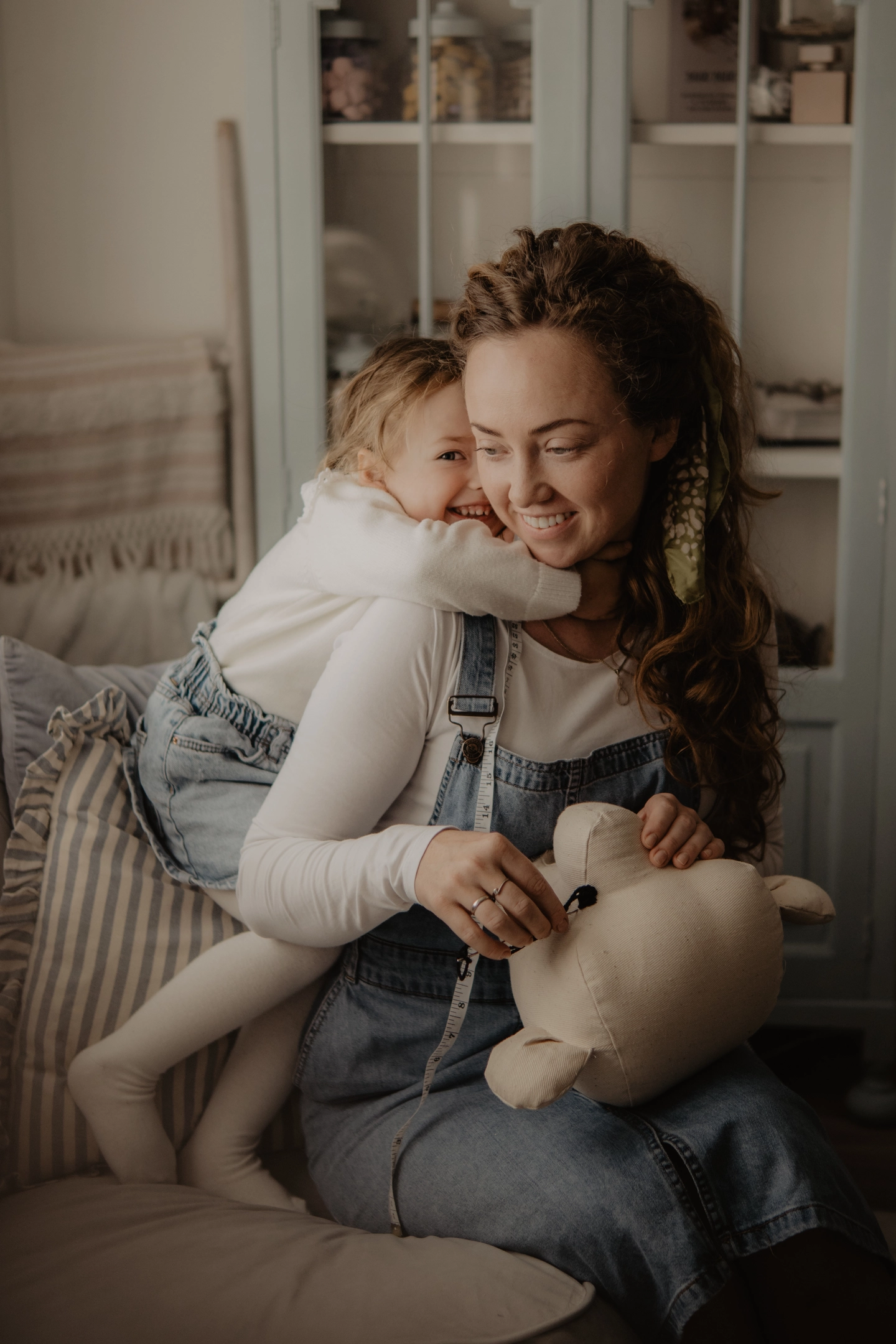A mother sewing a handmade decoration for a children’s room while her young daughter hugs her lovingly from the side, creating a warm and cozy moment of bonding and creativity.