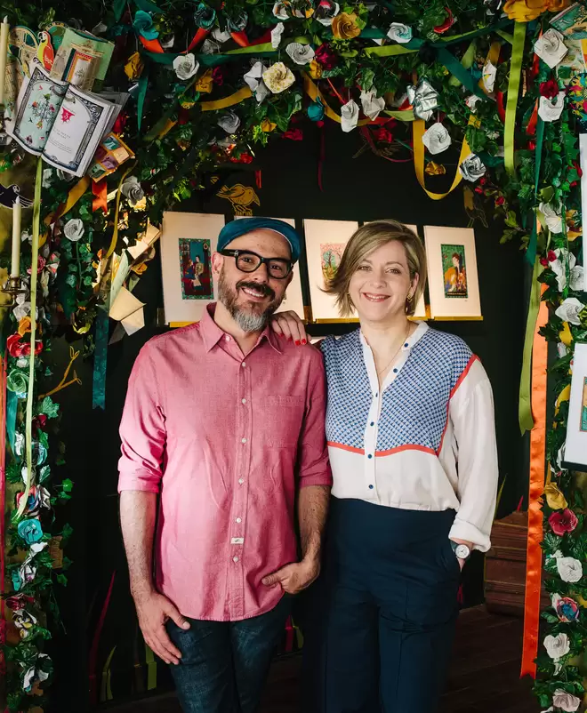 Eduardo Lima and Miraphora Mina, founders of MinaLima, smiling at the camera under a flower arch.
