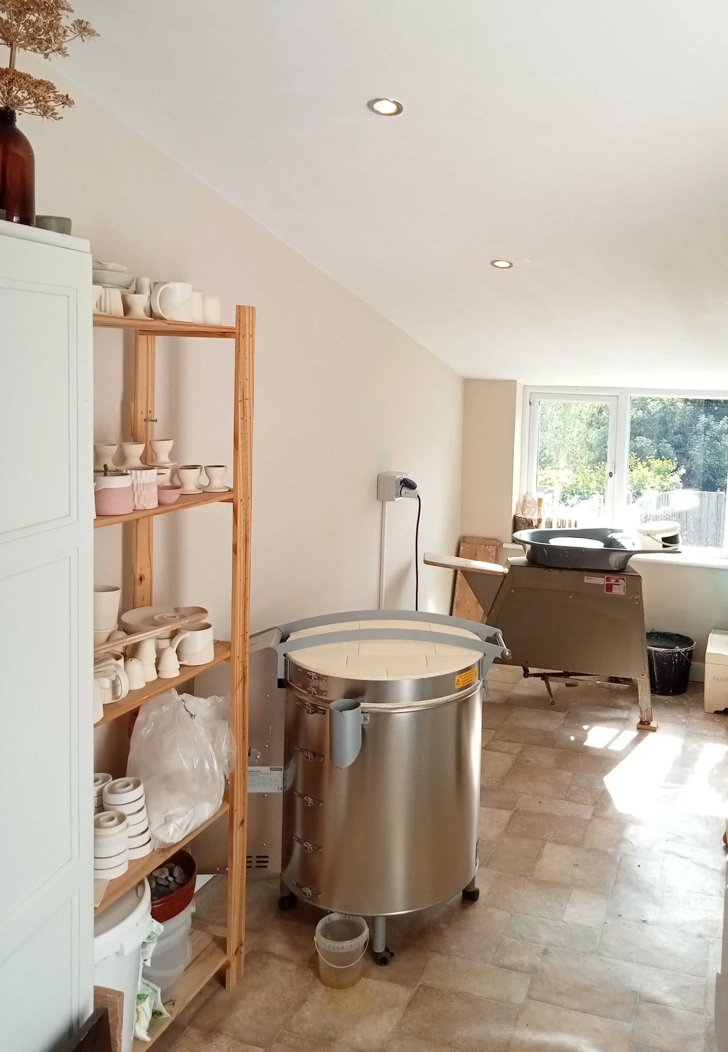 Faye Wellon Ceramics pottery studio in Herefordshire, Shows pottery wheel in the background, with the kiln and shelves of pottery in the foreground