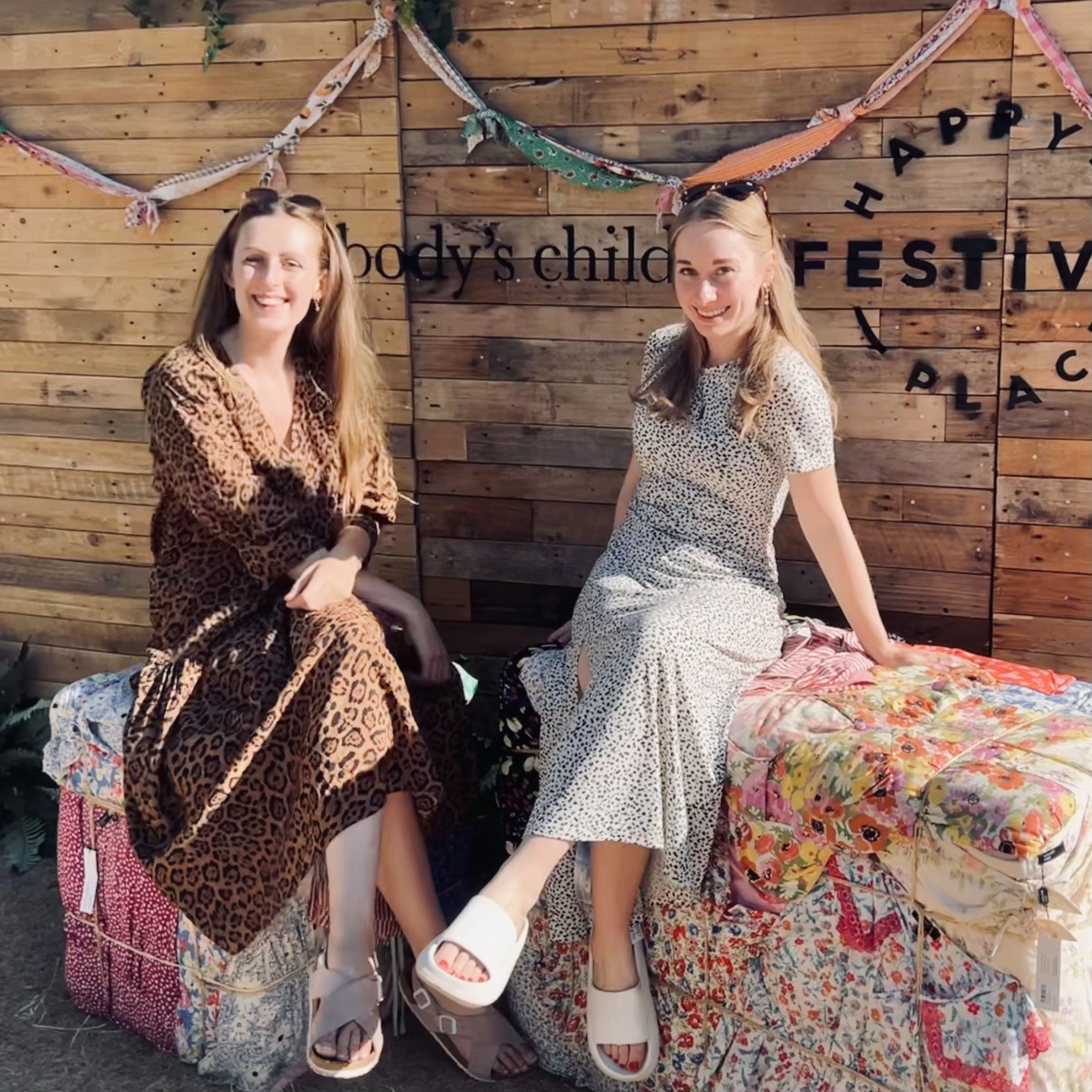 2 women sat on bales of hay covered in fabric, wearing long summer dresses