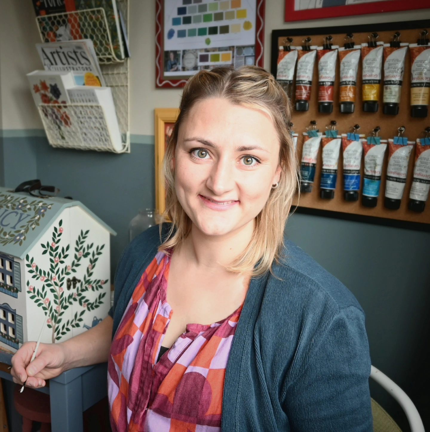 Caucasian woman with blonde hair sitting in an artist studio. She is smiling at the camera and has a small paintbrush in her hand. behind her is a dolls house that she is painting.  