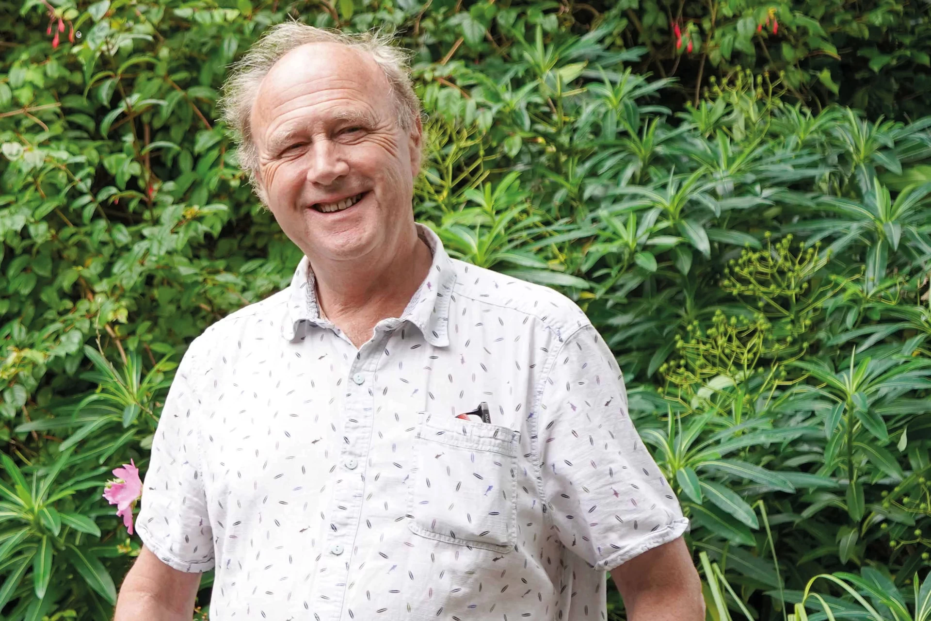 "Sir Tim Smit KBE, founder of The Eden Project, smiling at the camera, wearing a white  patterned shirt, stood infront of a green garden."