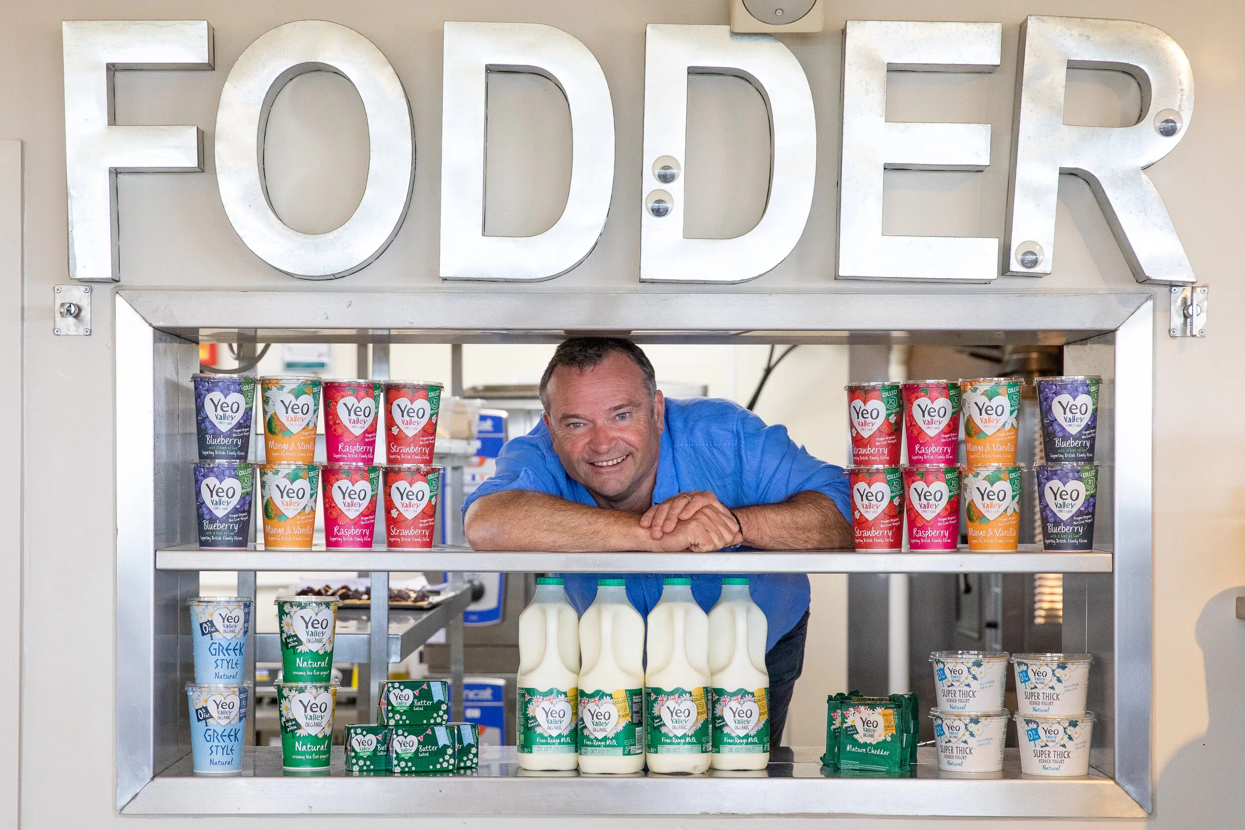 Tim Mead, founder of Yeo Valley, smiling at the camera, surrounded by Yeo Valley products, with a FODDER sign above him. 