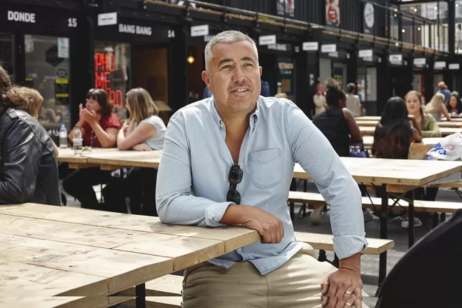 Roger Wade, founder Boxpark, smiling into the distance sat on a bench infront of Boxpark.