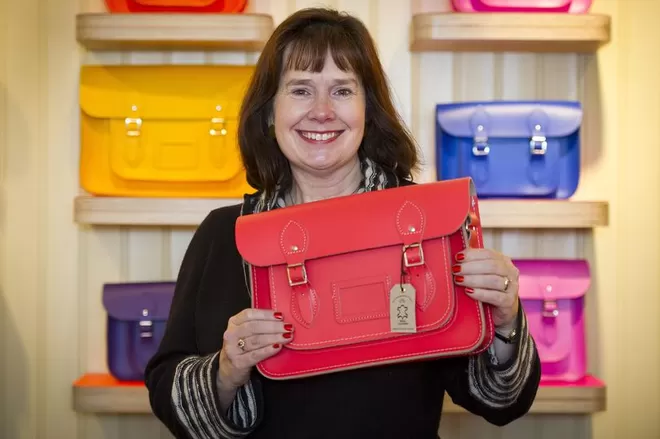 Julie Deane CBE, founder of The Cambridge Satchel Company, smiling at the camera holding a red satchel bag