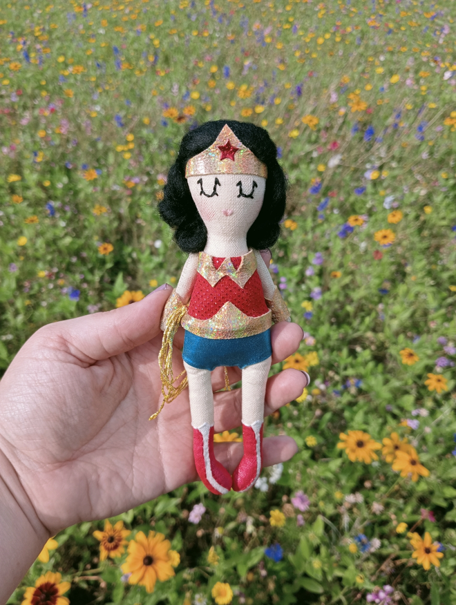 Mini decorative icon doll vintage style wonder woman, held in a hand to show scale. Wildflower background 