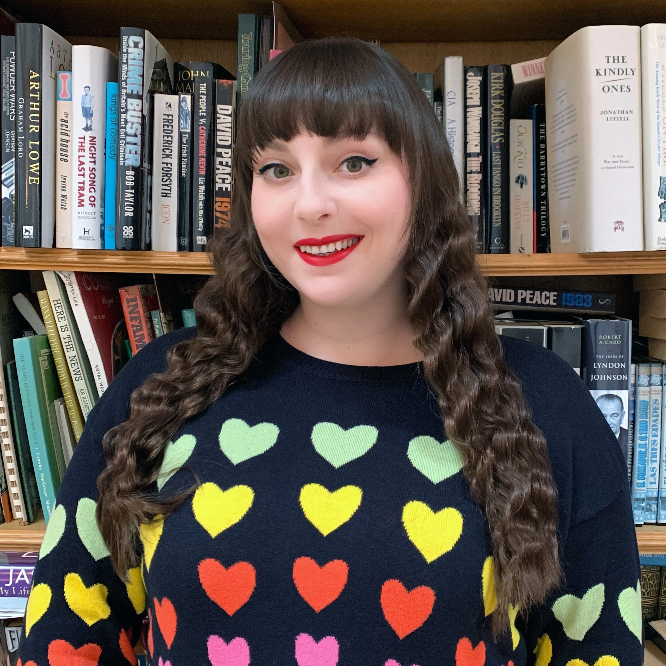 image shows a smiling woman with long brown hair. she is wearing red lipstick and her jumper has multicoloured hearts all over it.