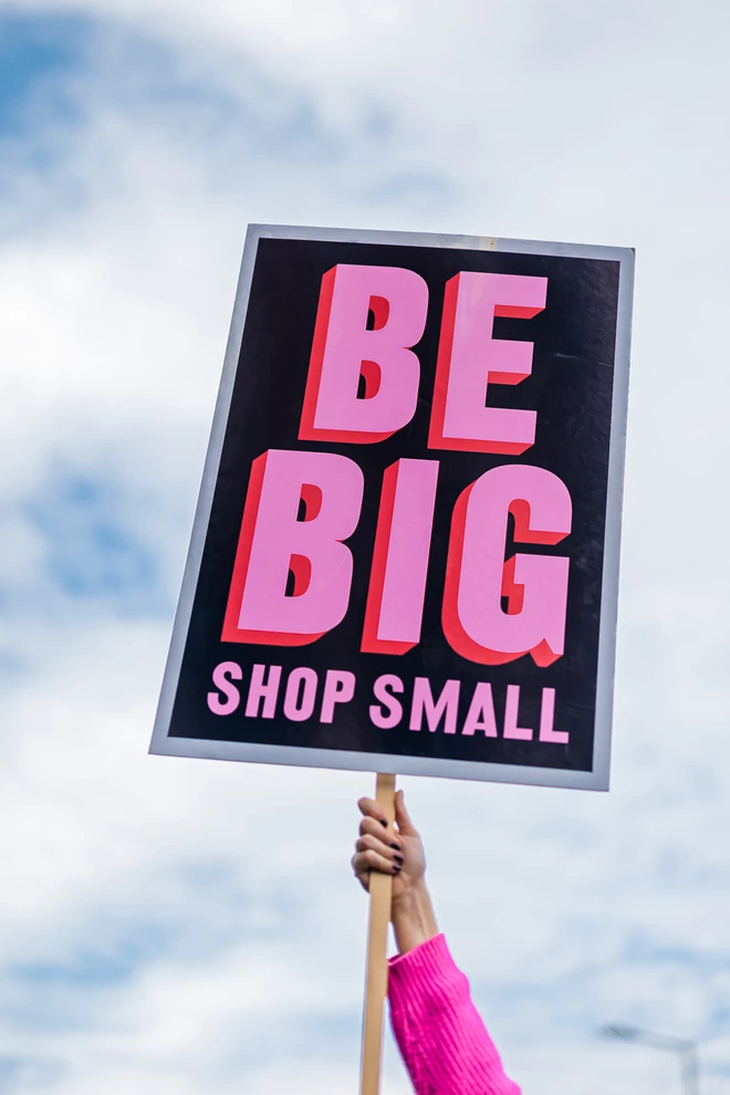 Be big shop small placard being held up with cloudy blue sky in the background