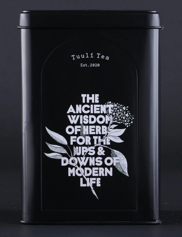 A black Tuuli Tea tin photographed on a grey background. It shows white text on a white flower graphic reading: THE ANCIENT WISDOM OF HERBS FOR THE UPS & DOWNS OF MODERN LIFE