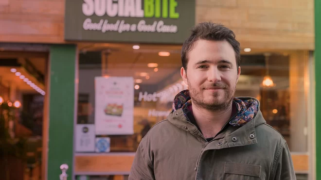 Josh Littlejohn, founder of The Social Bite, smiling at the camera, stood infront of a Social Bite shop.