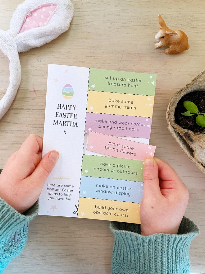A personalised Easter greetings card with seven cut out activity coupons is being held by a young child above a wooden desk.