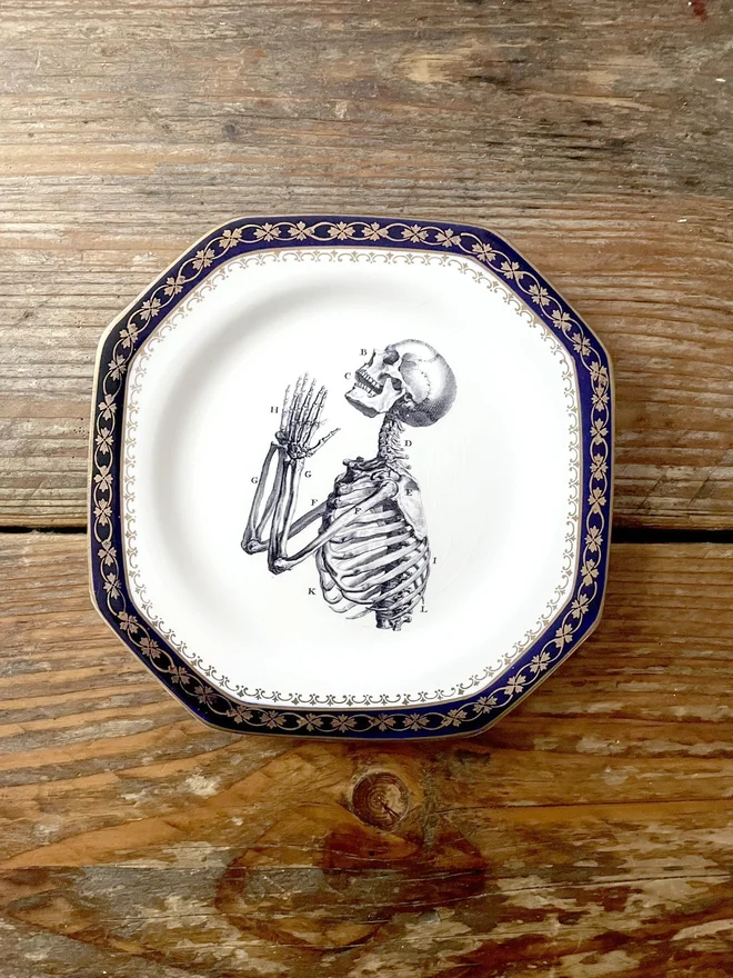 vintage plate with an ornate border, with a printed vintage illustration of a praying skeleton in the middle 