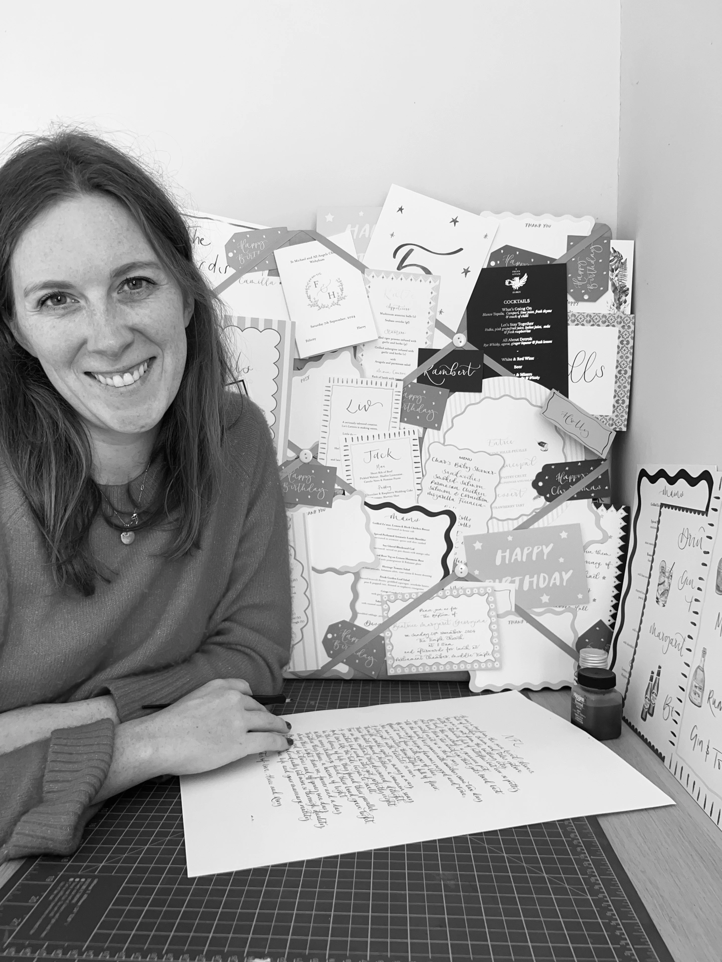 Liv sitting at her desk, with examples of her products displayed behind her. She is completing a calligraphy commission and smiling and the camera