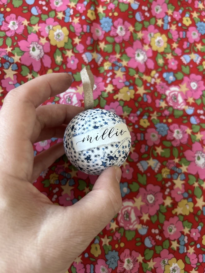 Mini Liberty fabric Christmas bauble with personalised Millie label held in front of a Liberty fabric backdrop 