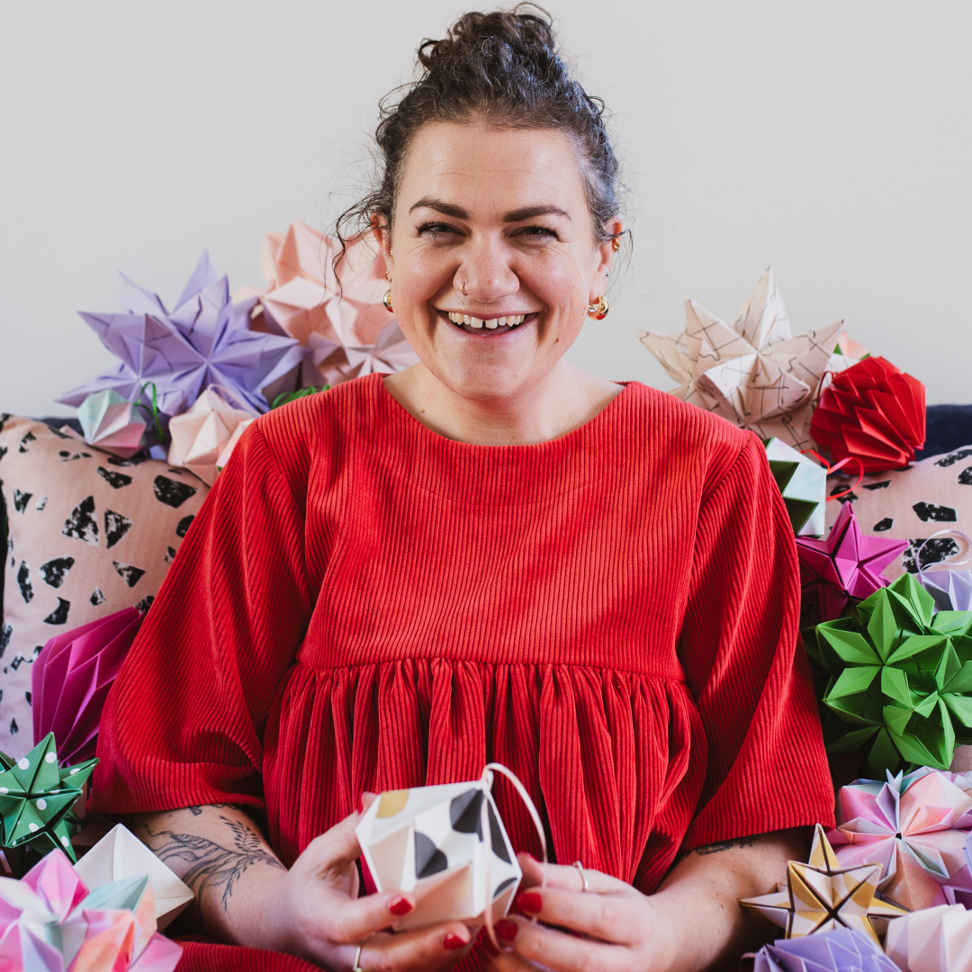 Woman in red dress sitting surrounded by colourful origami pieces.