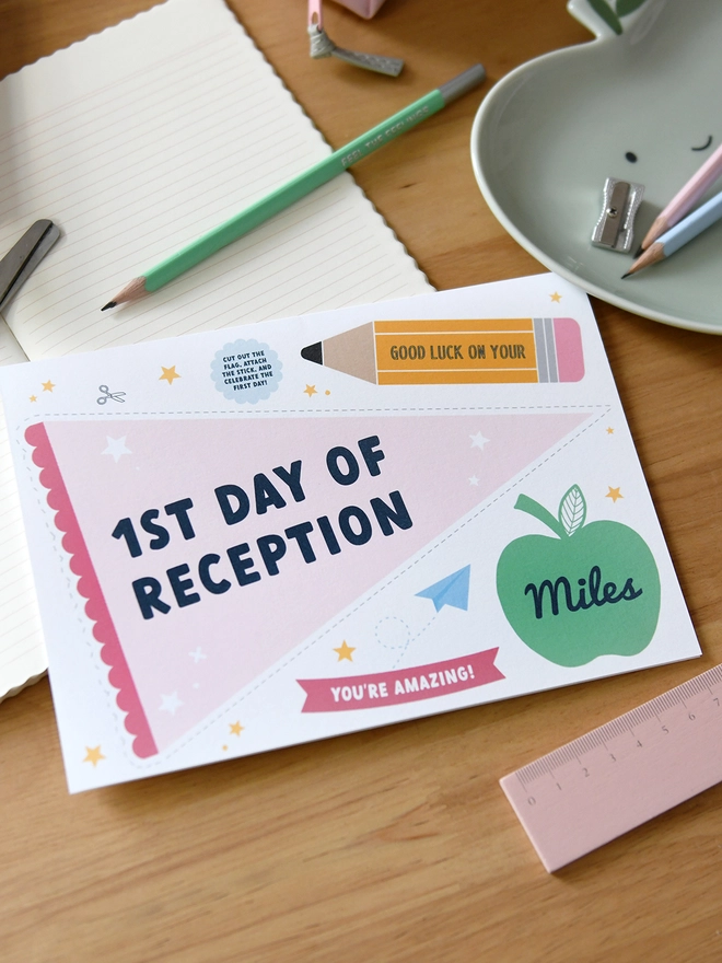  A "1st Day of Reception" card lies on a wooden desk surrounded by school supplies like scissors, pencils, a ruler, and a pencil case. The card includes a personalised name, encouraging messages, and playful designs.