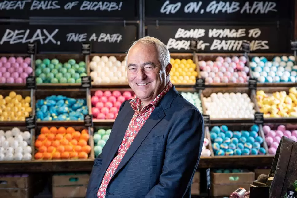 Mark Constantine, co-founder of Lush, smiling into the camera infront of a wall of Lush products.