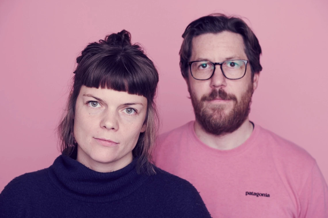Lucy Greenwood and Chris Renwick, founders of Lucy & Yak, looking into the camera, stood infront of a pink background.