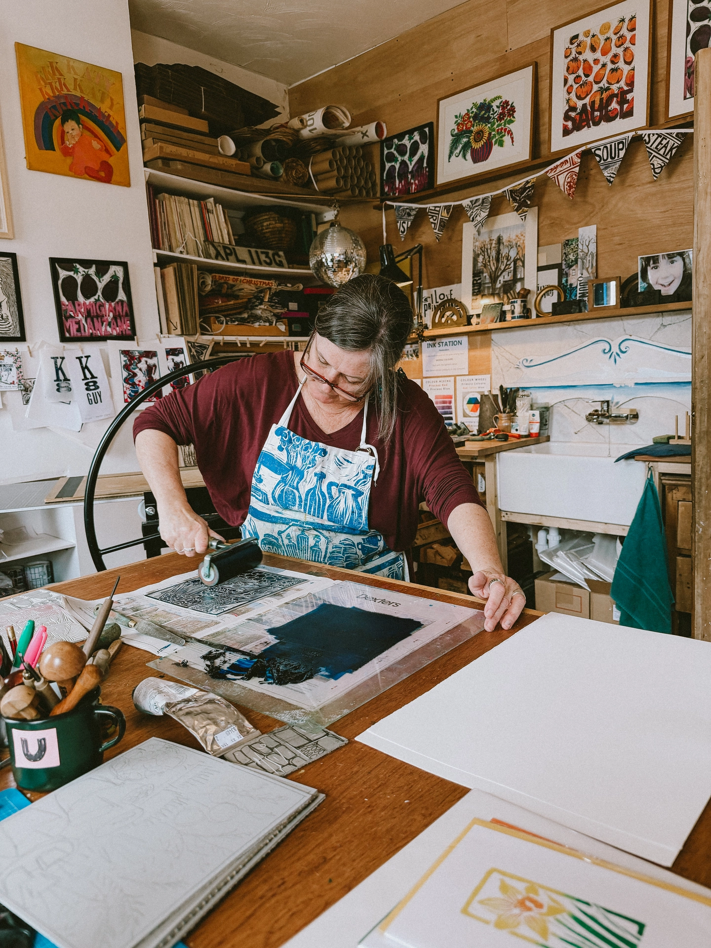 Printmaker Kate Guy in her London studio wearing a patterned blue apron, using a hand brayer roller to ink a linocut block on a wooden workbench surrounded by carving tools and artwork.