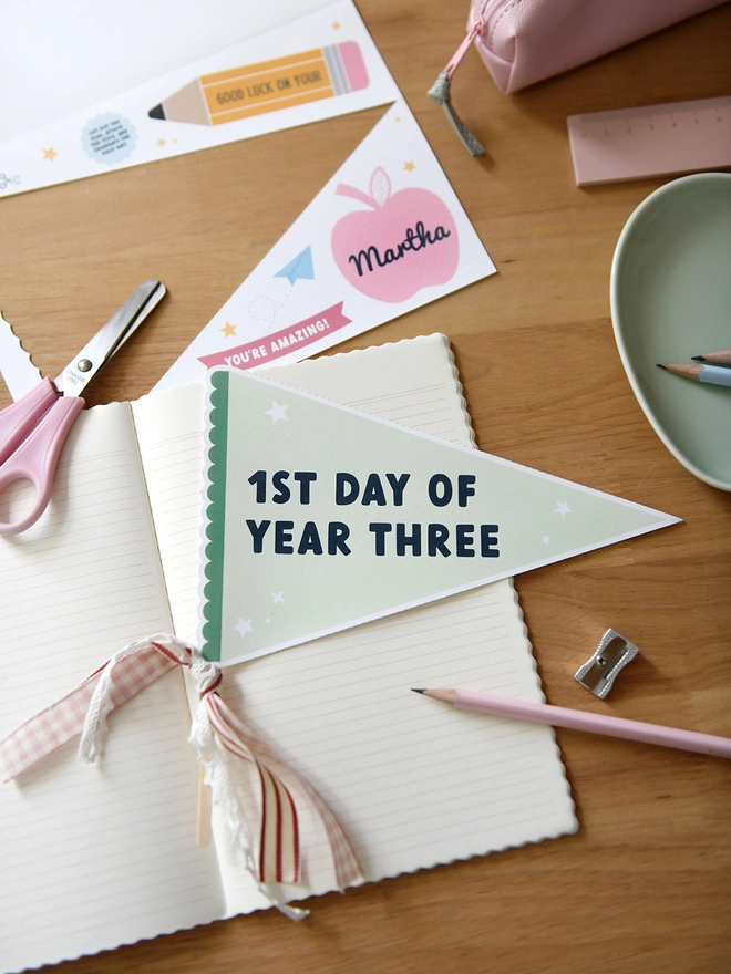 A green pennant flag with the words "1st Day Of Year Three" lays on an open notebook on a wooden desk. Various stationery items surround it.