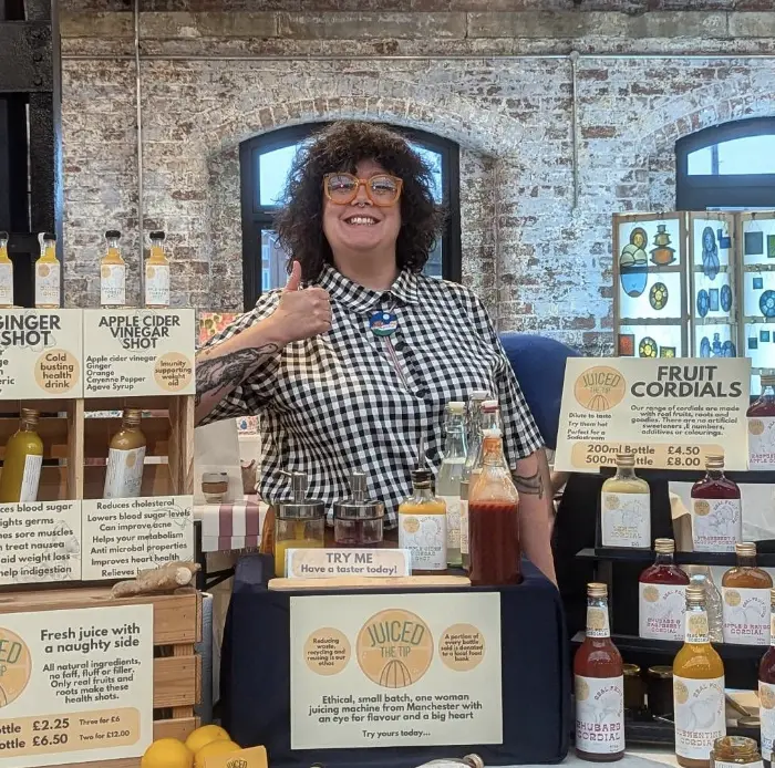 Katie stands behind a table at an event, wearing a checkered shirt and orange glasses. shes giving the thumbs up. On the table are her selection of wares and signs