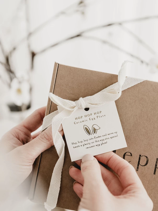 hand holding a gold foiled tag, tied around a brown gift box with cream ribbon