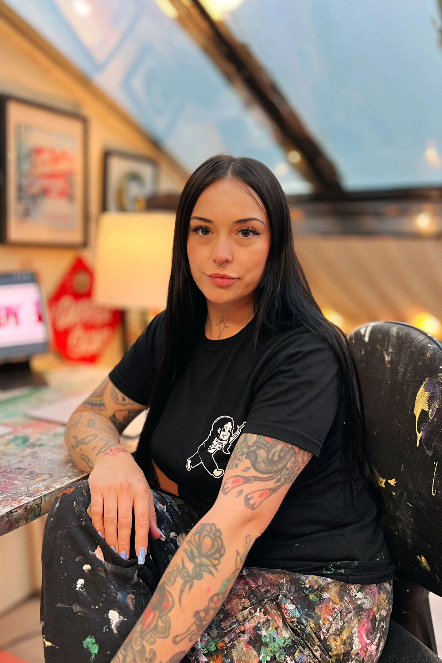 Tattooed woman with dark hair and black clothing with a lot of paint splatters. She is in a studio with paintings and photographson the wall.