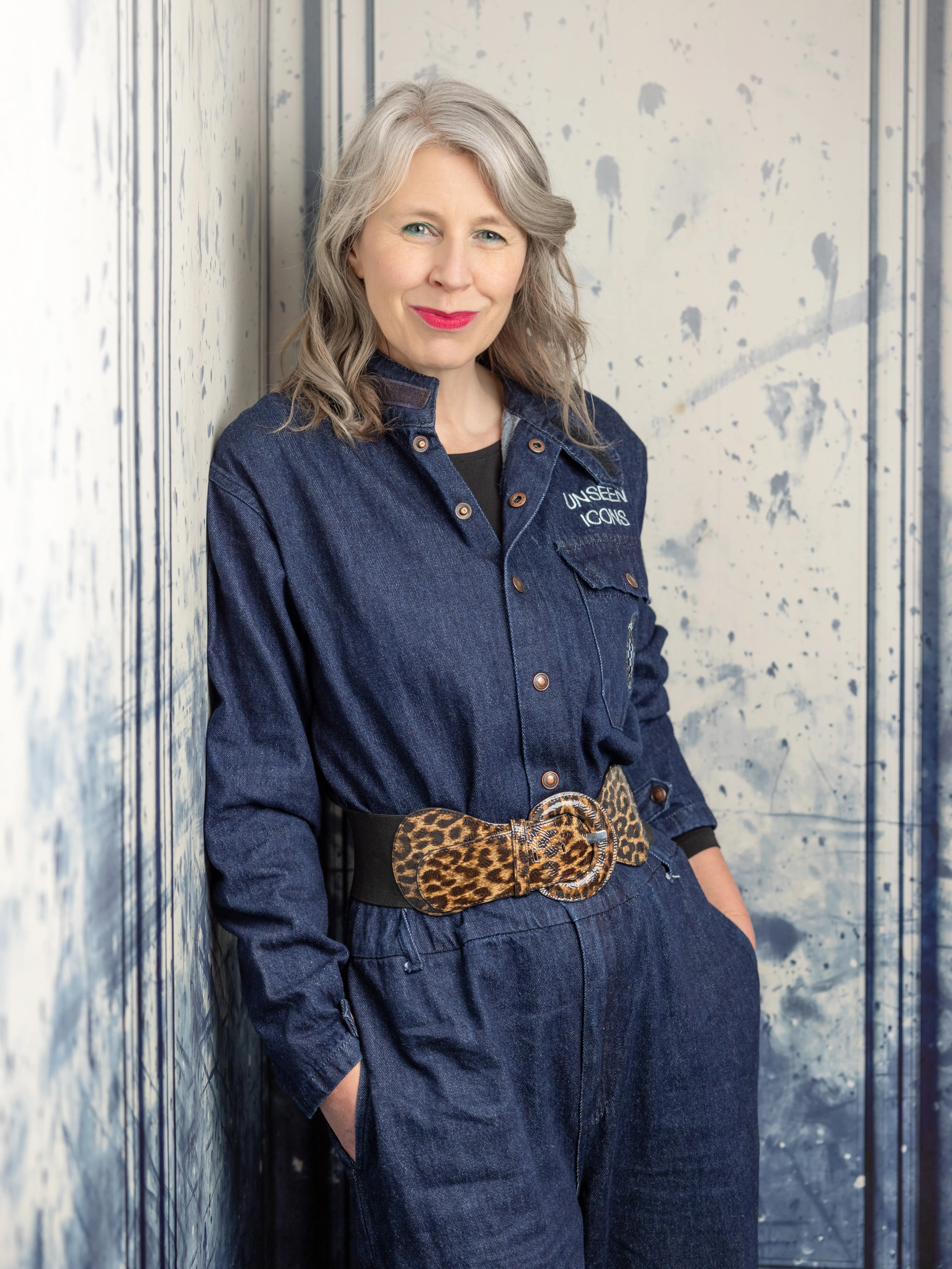 Unseen Icons founder Kerry Rutter leaning against a blue marbled wall in her studio wearing a denim boiler suit embroiled with Unseen Icons name