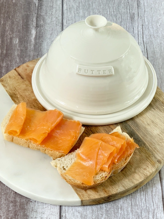 domed butter dish on circular wood and marble chopping board with smoked salmon on fresh bread