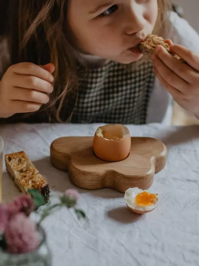 Wooden egg cloud cup with runny egg and toast soldiers with a little girl taking a bite of the toast