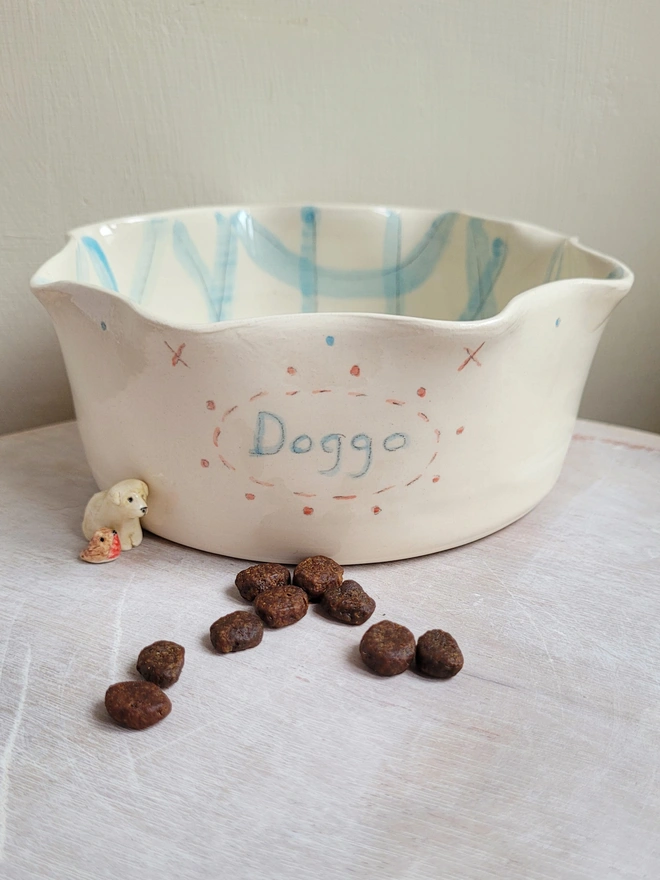 dog bowl held in a hand with the word doggo painted in blue with blue gingham check inside the dish there is a tiny dog figurine and robin figurine to the left of the bowl and small brown dog treats in front of it 