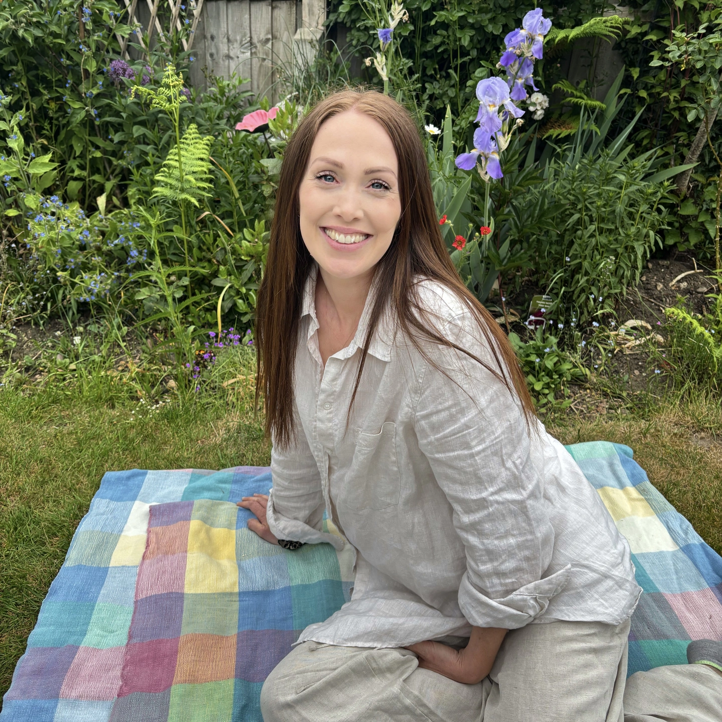 Girl sitting on patchwork rug outside in front of flowers and smiling at the camera