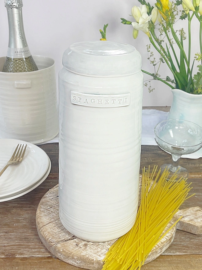 Spaghetti storage jar sat on a wooden table top amidst a scene of dried spaghetti, wine a jug of flowers and stack of plates