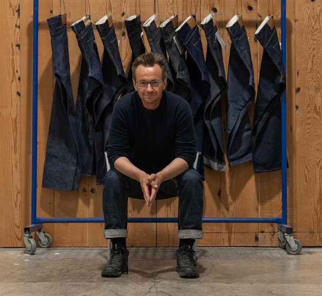 David Hieatt, founder of Howies, The Do Lectures & Hiut Denim, smiling at the camera, sat infront of a rack of Huit Denim jeans.