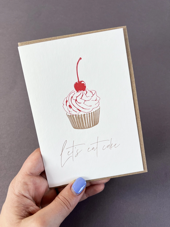 A little cupcake in a metallic gold cake case and a bright red cherry on top with "Let's eat cake" beautifully written in modern calligraphy underneath.