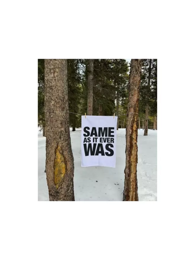 London Drying Same As It Ever Was white tea towel with black text hanging on twine in between 2 trees with trees in background and snow on ground