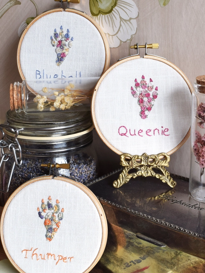 A display of 3 Rabbit Foot embroidery kits in their hoops, top to bottom are 1. Floral Meadow (Lavender Blues, Magenta & Buttermilk Yellow) 2. Pink Roses (5 shades of pink).  3. Sunshine Garden (Oranges, Yellows, Purples).