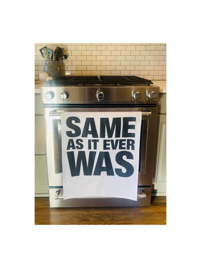 London Drying Same As It Ever Was white tea towel with black text hanging on stainless steel oven door with comtainer of kitchen utensils on counter above
