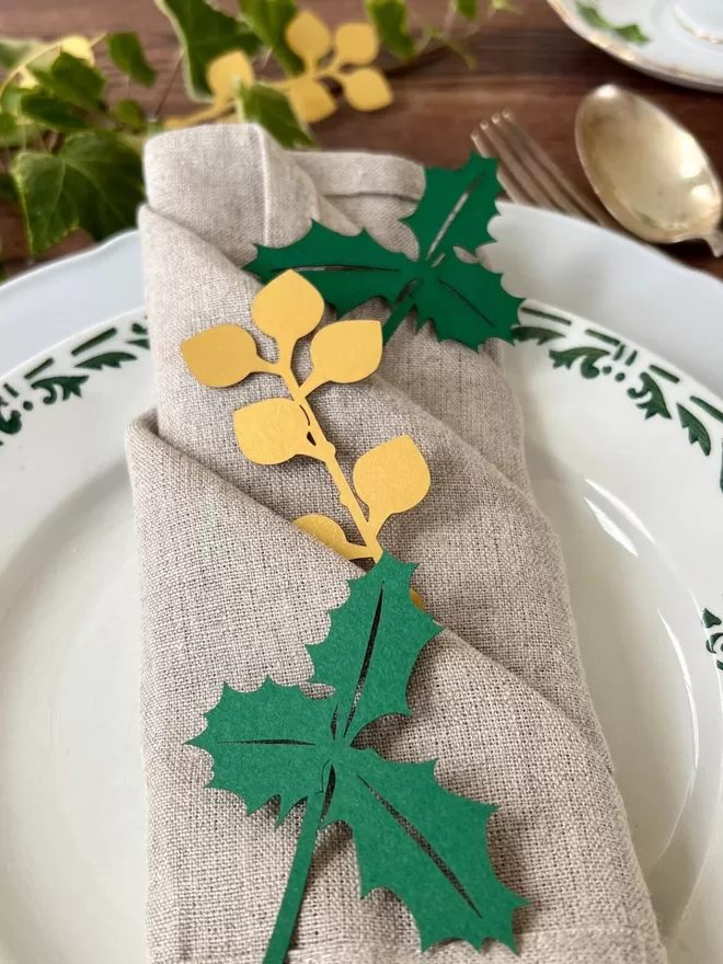 Folded linen napkin with green paper holly sprigs and gold paper laurel berry decoration placed into folds of napkinWinter Foliage Holiday Place Setting