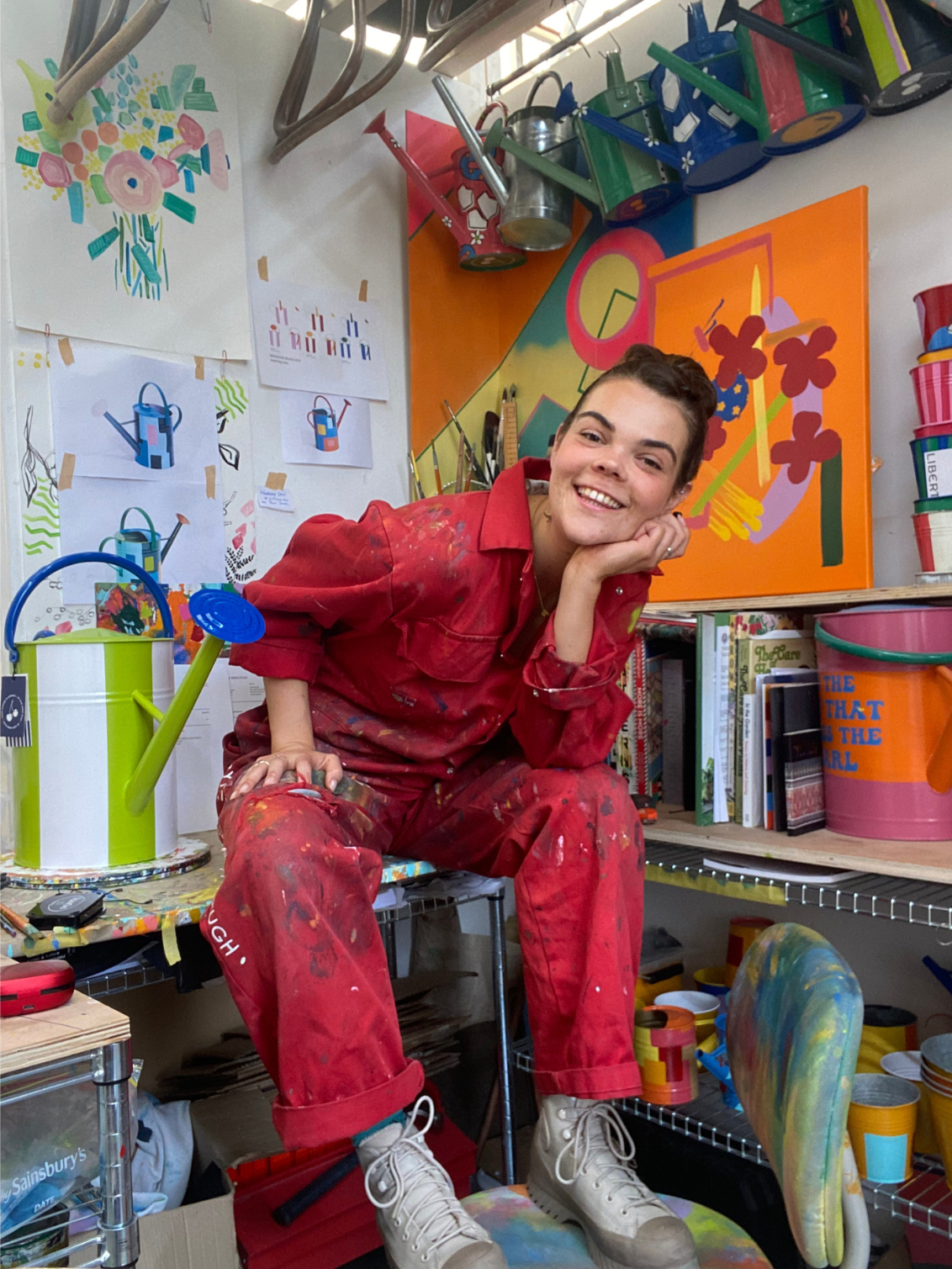Photo of artist Julie-Anne Pugh, sat on her desk in the colourful studio in South East London with hand painted watering cans surrounding her.
