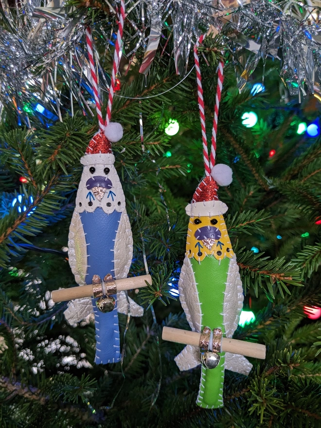 Two hand stitched faux leather budgie Christmas decorations wearing Santa hats. One bird is blue, one green.