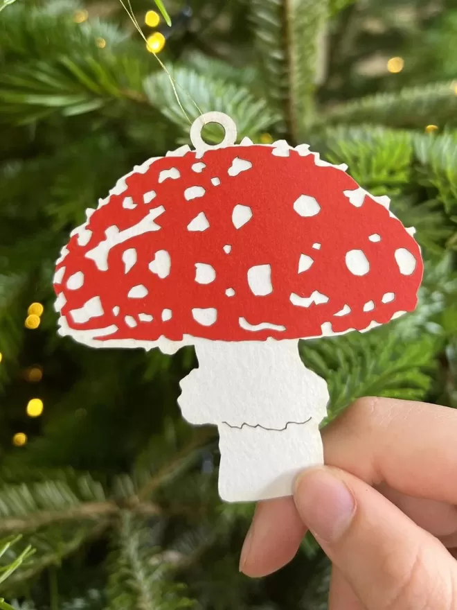 Christmas Mushroom Ornament with bright red cap with white spots.  Mushroom has a short wide white stalk.  Ornament is hanging on a Christmas tree.