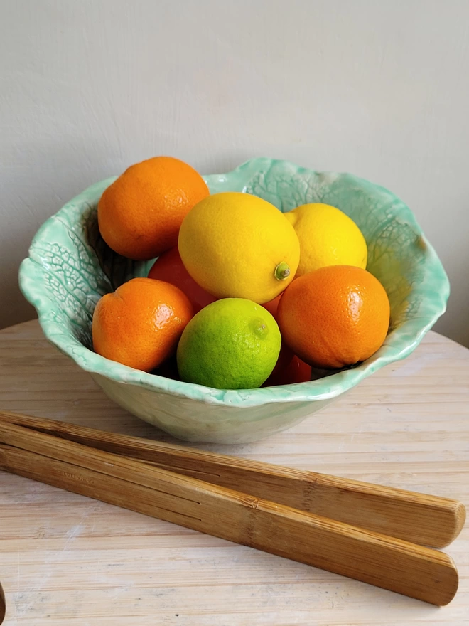 a green glazed ceramic cabbage leaves fruit bowl with oranges, lemons and a lime inside and some bamboo servers in the foreground
