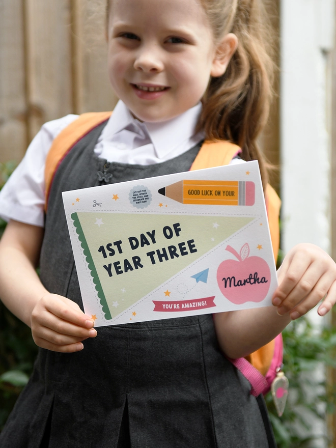 A young child, wearing a school uniform and backpack, holds a "1st Day of Year Three" card with excitement. The card features encouraging messages and playful designs, perfect for celebrating a child's first day at school.